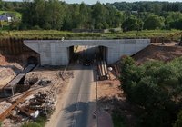 Z perspektywy lotu ptaka widok na nowo budowany wiadukt drogowy pod linią kolejową w Łącznej. Widoczne są betonowa konstrukcja obiektu, przebudowywany układ drogowy z ruchem samochodowym, wykopy, umocnienia oraz sprzęt budowlany.