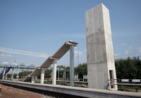 Z perspektywy centralnej widok na budowaną kładkę dla pieszych od strony peronu. Widoczne są żelbetowy szyb windy, konstrukcja schodów, nowy peron oraz pracownik prowadzący roboty budowlane.